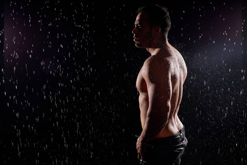 Przystojny mężczyzna z umięśnionym ciałem, pozujący w deszczu, z nagim tułowiem, kroplami wody na ciele - Zdjęcie, obraz