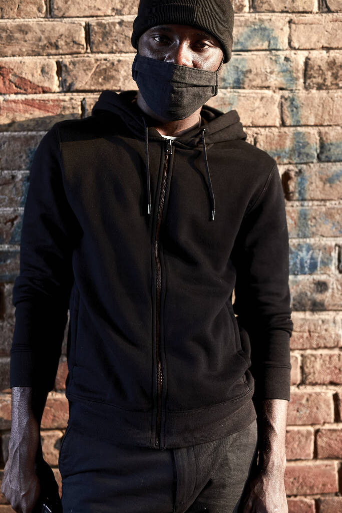 Retrato do jovem homem africano mascarado em pé junto à parede de tijolos, hooligan vestido de preto - Foto, Imagem