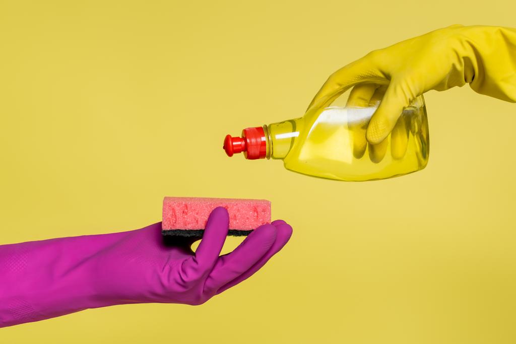 обрезанный вид людей в резиновых перчатках, держащих бутылку с моющим средством и губку изолированы на желтый  - Фото, изображение