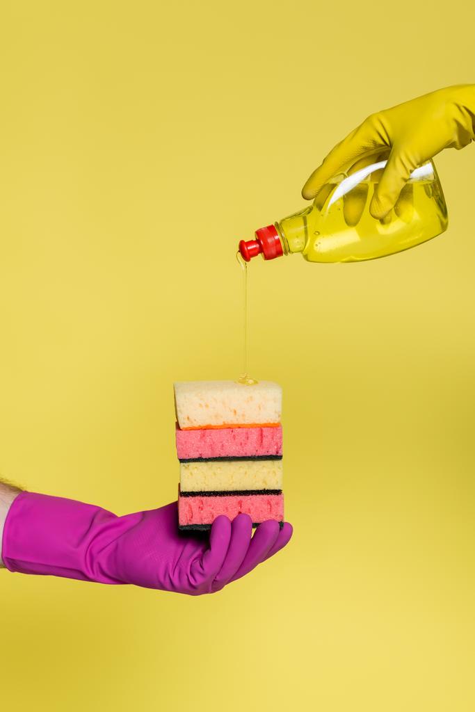visão recortada de pessoas em luvas de borracha segurando garrafa com detergente e pilha de esponjas isoladas em amarelo  - Foto, Imagem