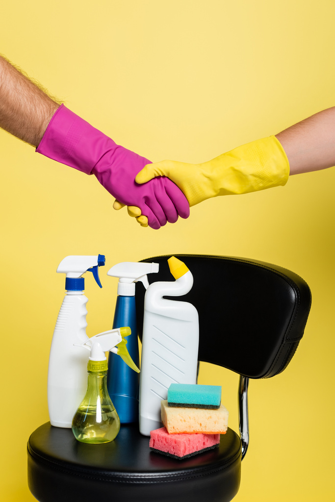 vista recortada de los limpiadores estrechando las manos cerca de llegar a las botellas con detergente y esponjas limpias en la silla aislada en amarillo - Foto, imagen