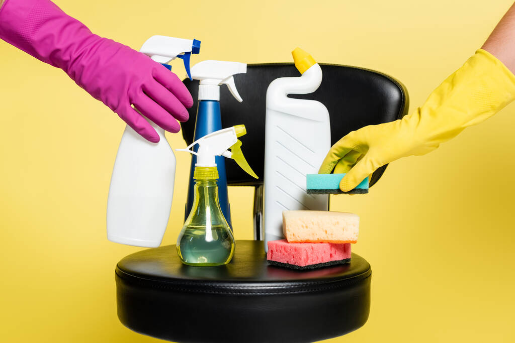 vue recadrée des nettoyants atteignant bouteille avec détergent et éponge sur chaise noire isolée sur jaune - Photo, image