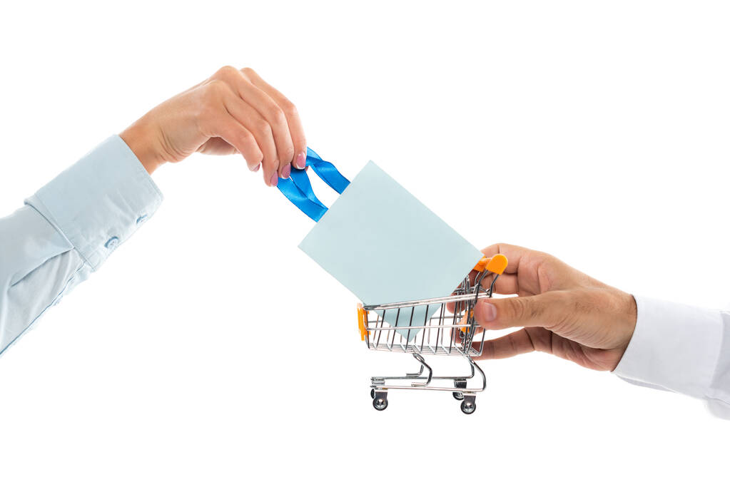 vista recortada del hombre sosteniendo pequeño carrito de compras cerca de la mujer con bolsa de papel aislado en blanco  - Foto, Imagen