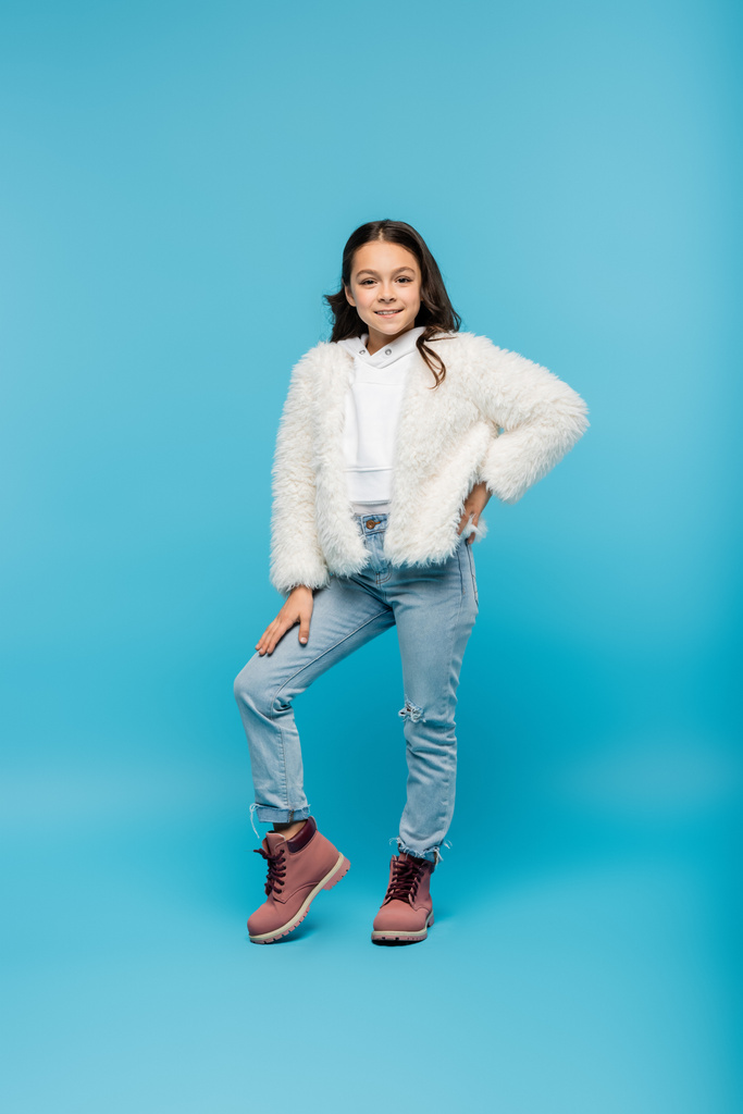 longitud completa de niña preadolescente positiva en chaqueta de piel sintética y botas de invierno posando con la mano en la cadera en azul - Foto, imagen