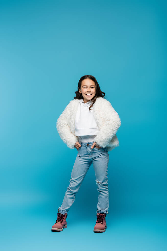 longitud completa de la niña preadolescente feliz en chaqueta de piel sintética y botas de invierno de pie con las manos en los bolsillos en azul - Foto, imagen