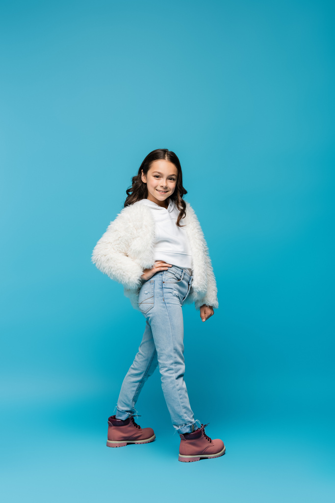 full length of smiling preteen girl in faux fur jacket and winter boots standing with hand on hip on blue - Photo, Image