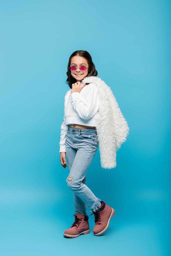 pleine longueur de jeune fille heureuse en lunettes de soleil rose tenant veste blanche en fausse fourrure sur bleu - Photo, image