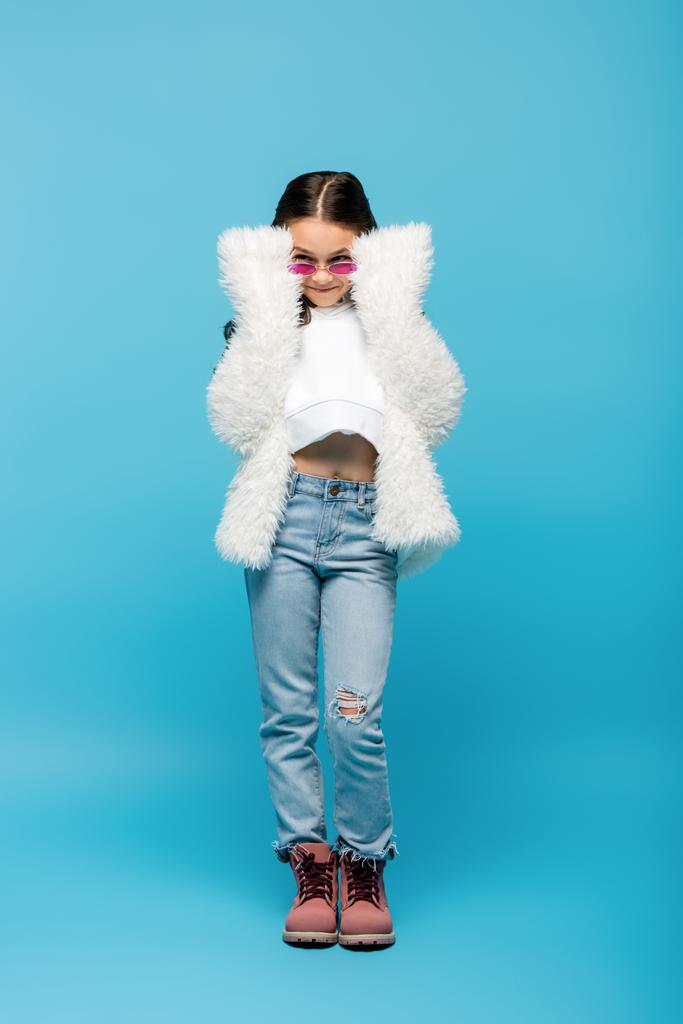 longitud completa de la niña preadolescente sonriente en gafas de sol de color rosa y chaqueta de piel sintética blanca en azul - Foto, imagen
