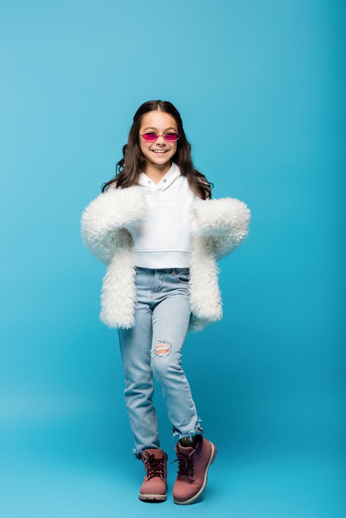 longitud completa de la niña preadolescente sonriente en gafas de sol de color rosa y chaqueta de piel sintética blanca posando en azul - Foto, imagen