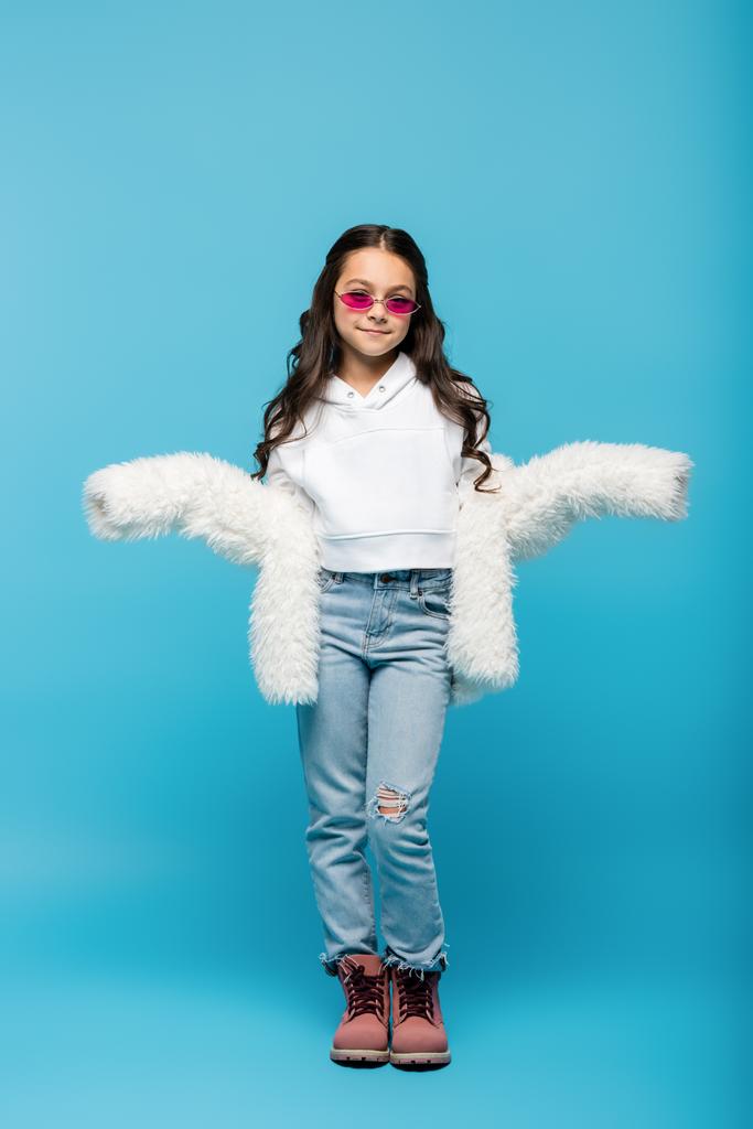 longitud completa de la niña preadolescente sonriente en gafas de sol de color rosa y chaqueta de piel sintética blanca que muestra gesto de encogimiento de hombros en azul - Foto, Imagen