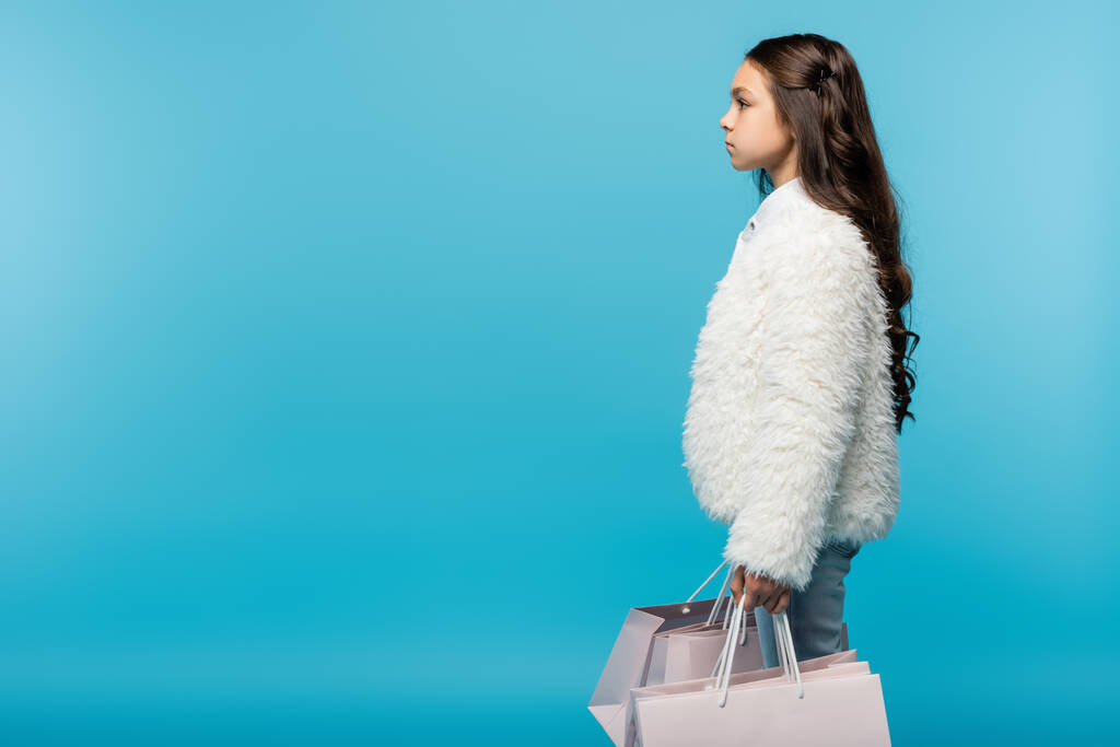 vista lateral de niña preadolescente en chaqueta de piel sintética blanca sosteniendo bolsas de compras en azul - Foto, Imagen