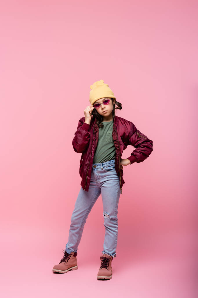полная длина девочки-подростка в модном наряде регулируя солнцезащитные очки и позируя с рукой на бедре на розовый  - Фото, изображение