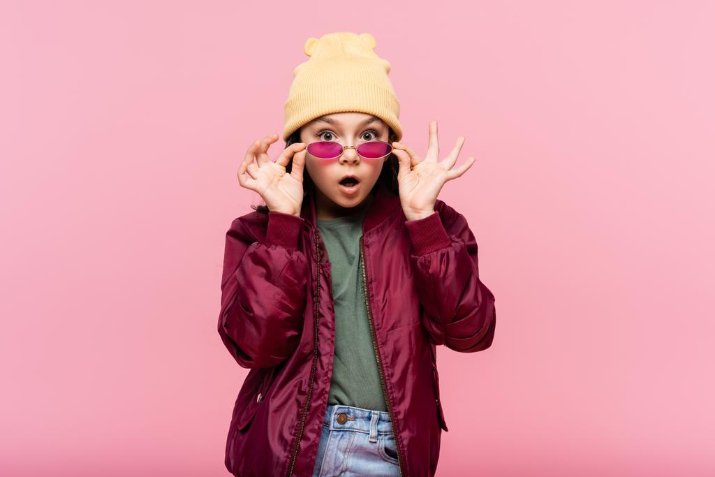 verrast tiener meisje in trendy outfit aanpassen zonnebril geïsoleerd op roze - Foto, afbeelding