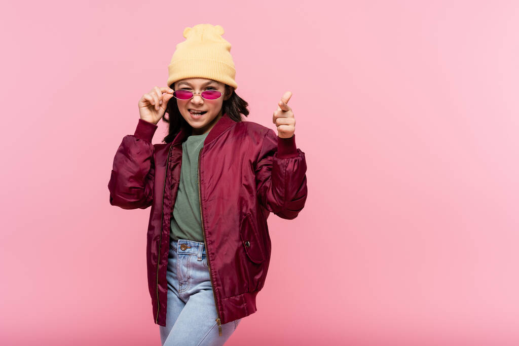 happy preteen girl in trendy outfit adjusting sunglasses and pointing at camera isolated on pink - Photo, Image