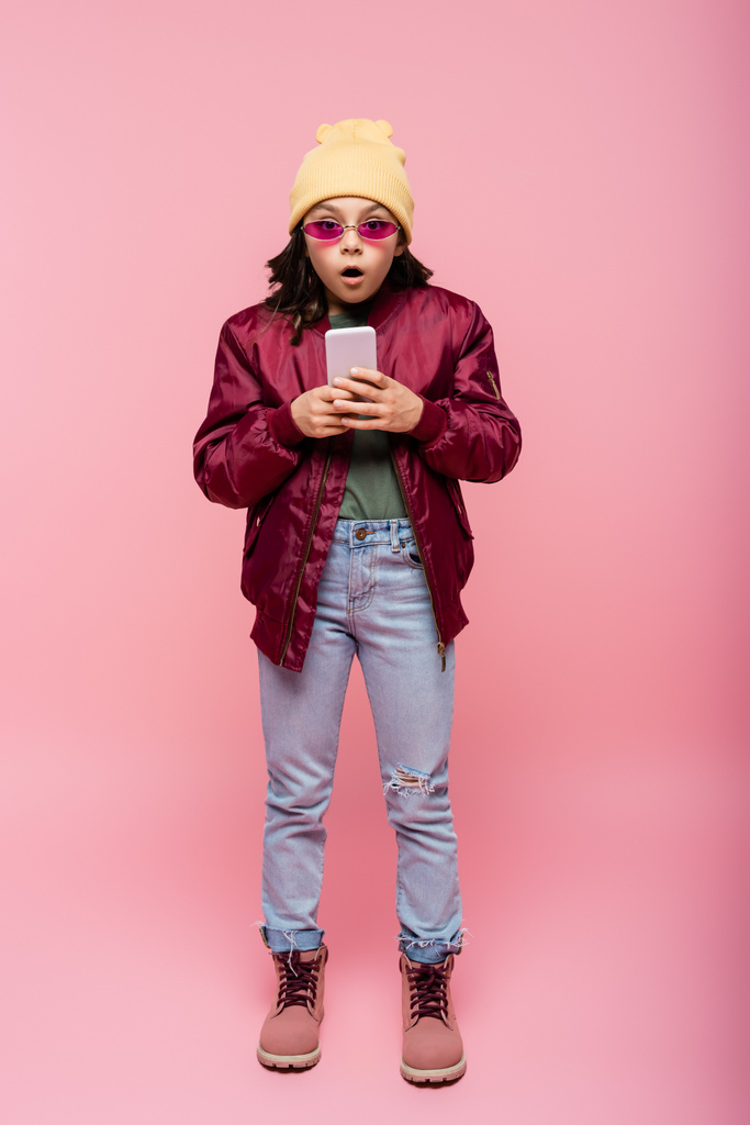 piena lunghezza di scioccato preteen girl in abito alla moda utilizzando smartphone su rosa  - Foto, immagini