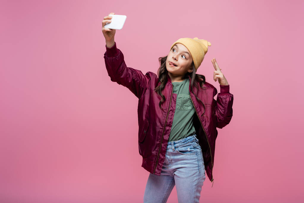 nastolatka dziewczyna w stylowy strój biorąc selfie i pokazując znak pokoju izolowane na różowy  - Zdjęcie, obraz