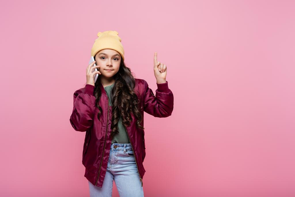 tiener in stijlvolle outfit praten op mobiele telefoon en wijzen met vinger geïsoleerd op roze  - Foto, afbeelding