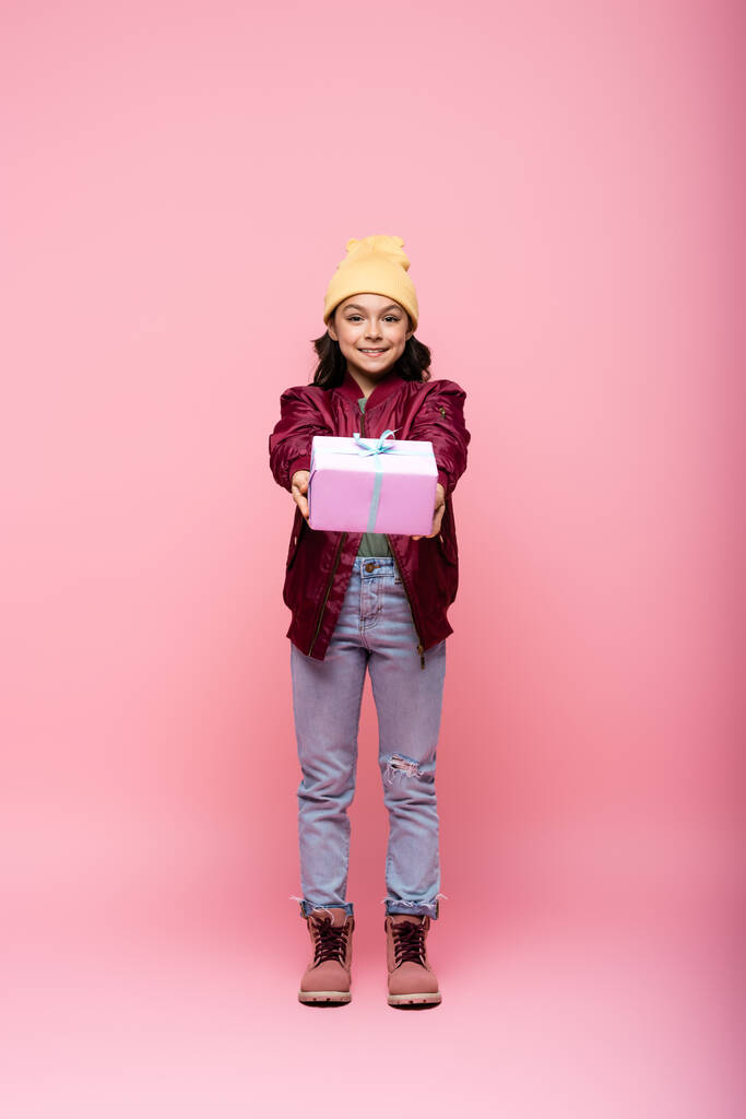 volle Länge der glücklichen preteen Mädchen im Winter-Outfit hält eingewickelte Geschenk auf rosa  - Foto, Bild
