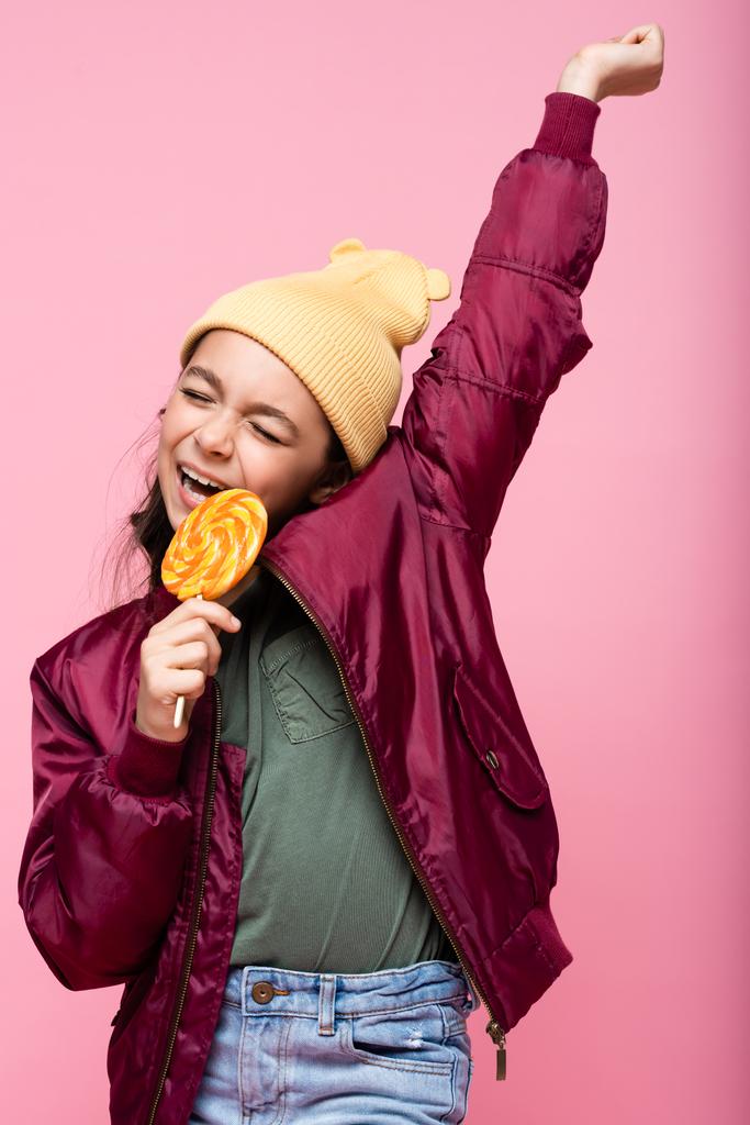 cheerful preteen girl in winter outfit holding lollipop and smiling isolated on pink  - Photo, Image