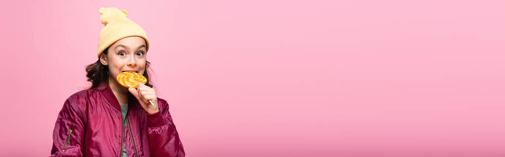 stylový preteen dívka v zimním oblečení kousání lízátko izolované na růžové, banner - Fotografie, Obrázek