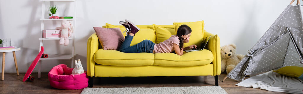 девочка-подросток лежит на диване и пользуется ноутбуком в современной гостиной, баннер - Фото, изображение