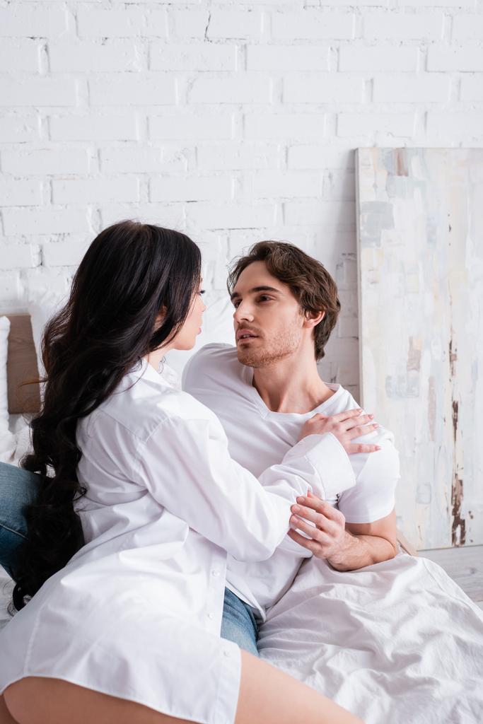 молодая и сексуальная пара в белой одежде смотрит друг на друга, обнимаясь в спальне - Фото, изображение