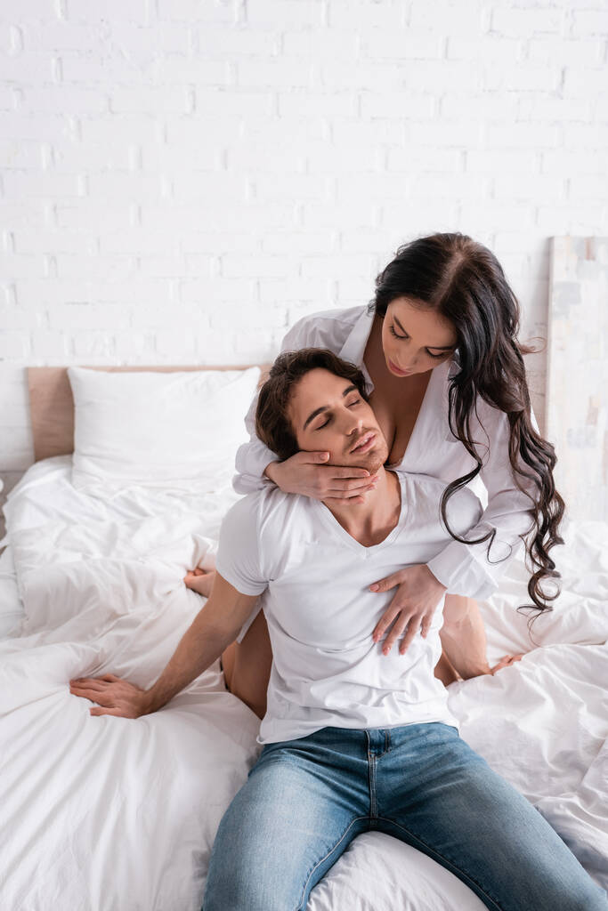 sexy vrouw verleiden jonge man zitten met gesloten ogen in slaapkamer - Foto, afbeelding