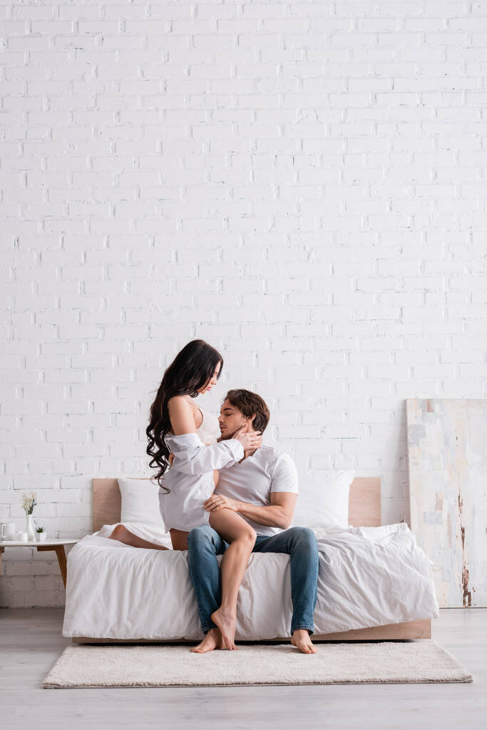 vrouw in wit shirt knuffelen vriendje zitten op bed in jeans - Foto, afbeelding