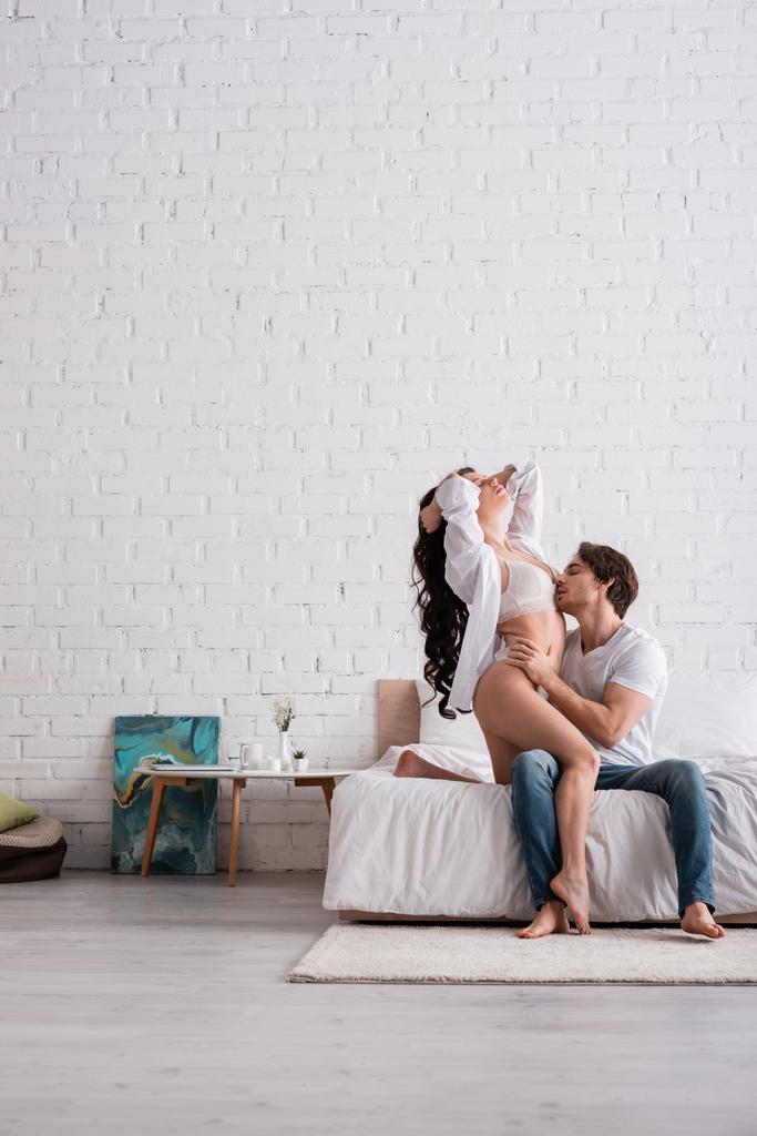 man in jeans embracing passionate woman in lingerie and white shirt in bedroom - Photo, Image