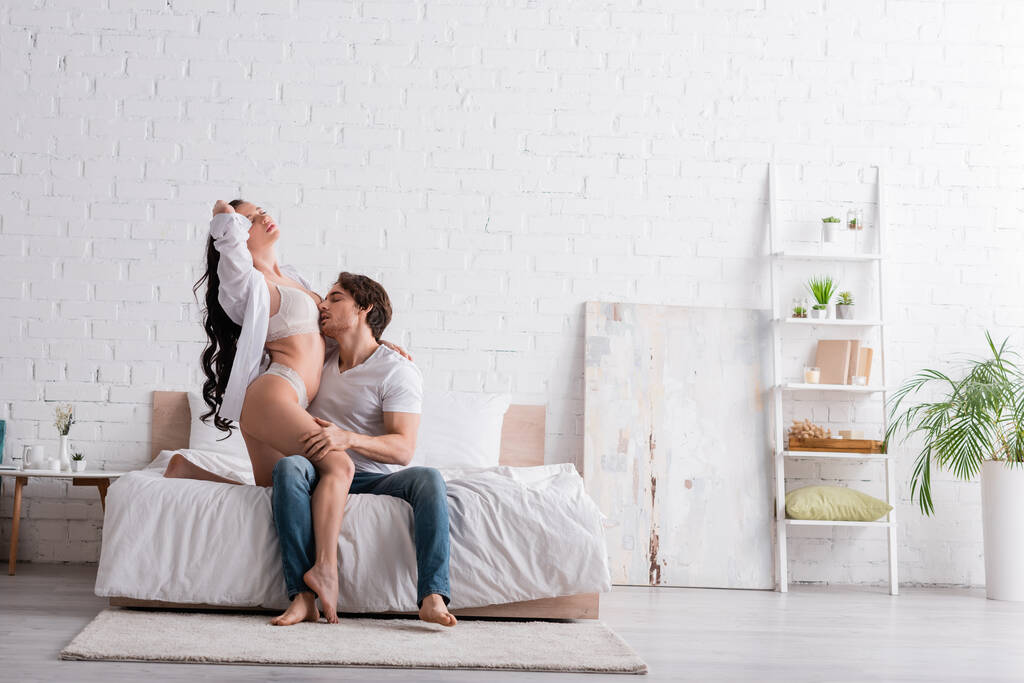femme séduisante en lingerie et chemise blanche près de l'homme l'embrassant dans une chambre spacieuse - Photo, image