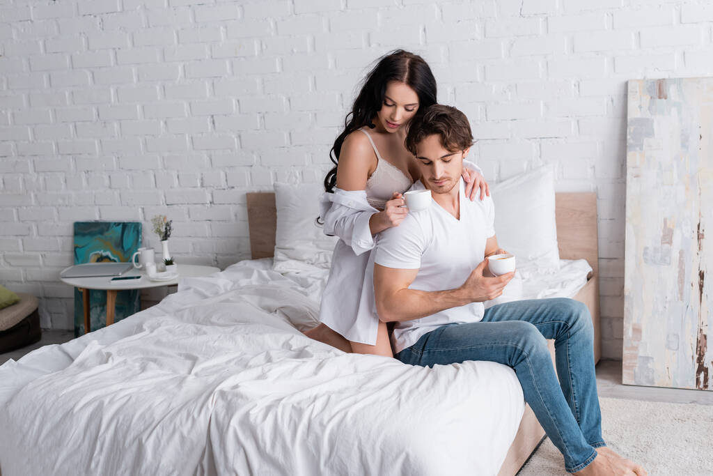страстная женщина в рубашке и лифчике соблазняет мужчину, когда пьет кофе в спальне - Фото, изображение