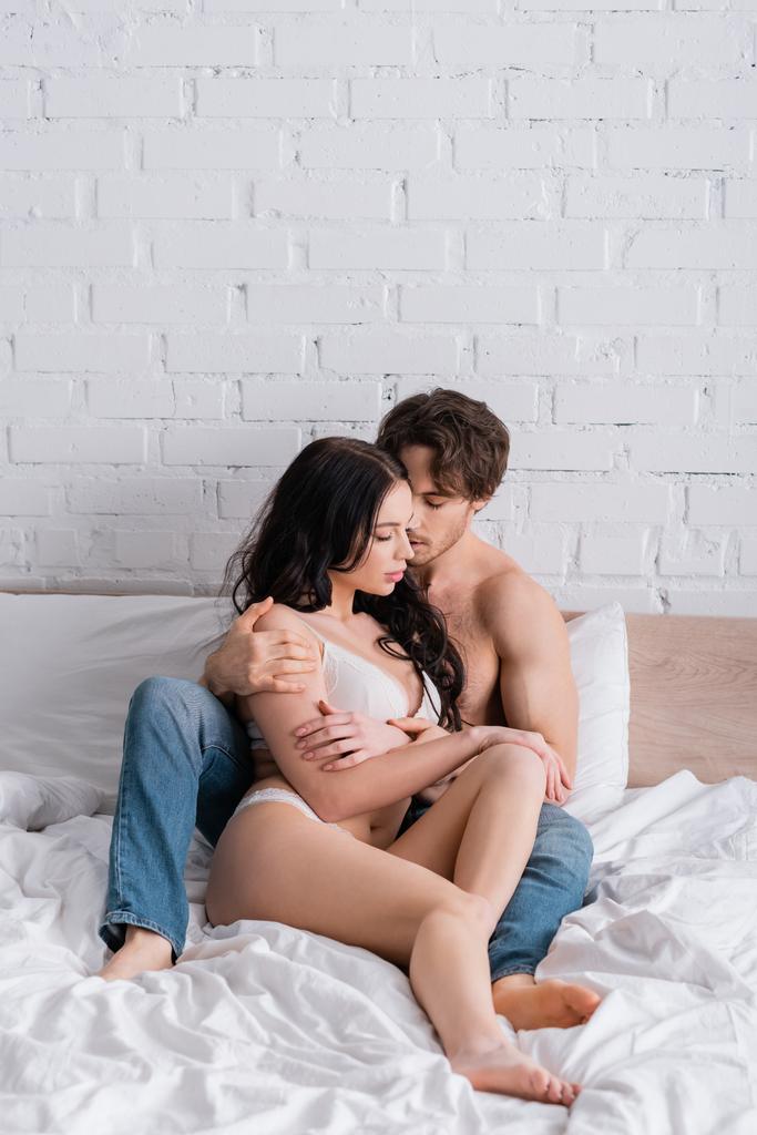 sexy man in jeans hugging woman in lingerie while sitting on bed with closed eyes - Photo, Image
