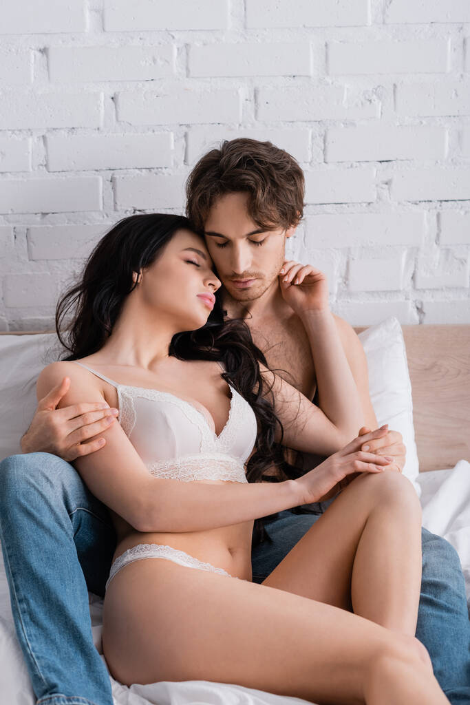 vášnivá žena ve spodním prádle a bez trička muž v džínách objímání na posteli se zavřenýma očima - Fotografie, Obrázek
