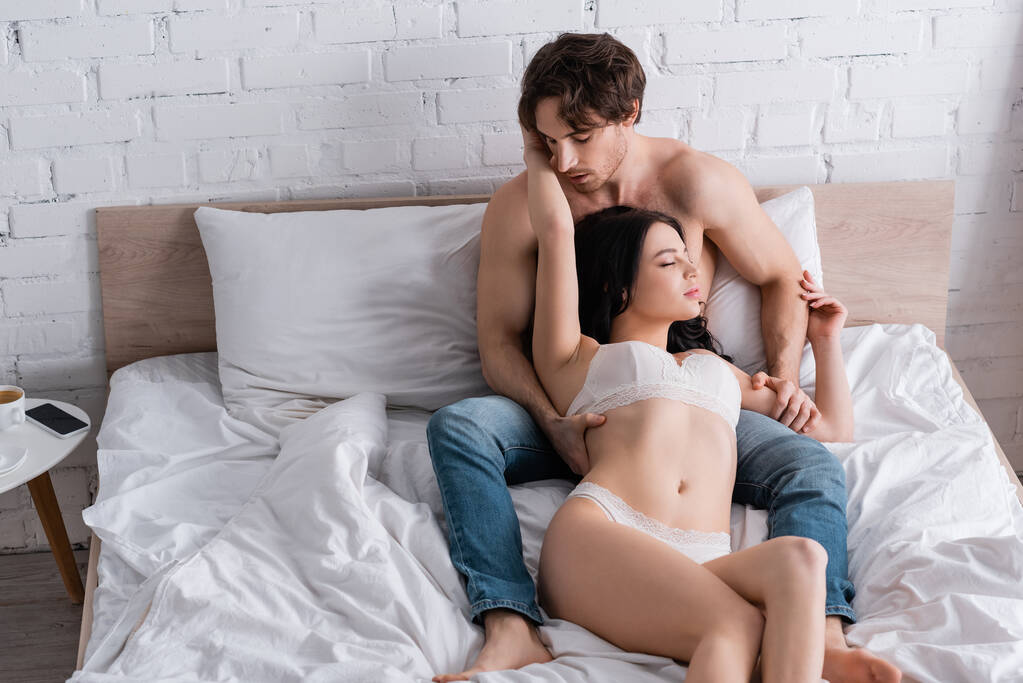 shirtless man in jeans and passionate woman in lingerie embracing on white bedding - Photo, Image