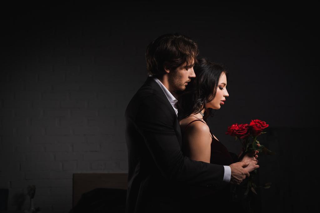 Seitenansicht eines Mannes im Anzug neben einer sinnlichen brünetten Frau mit roten Rosen  - Foto, Bild