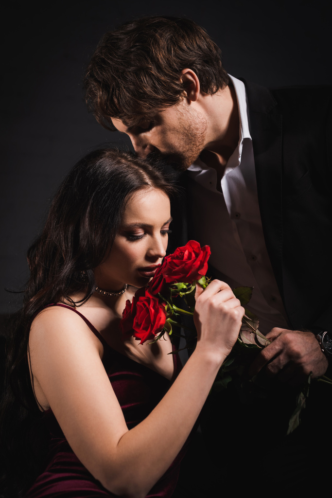 elegante Frau riecht rote Rosen in der Nähe eines Mannes im Anzug auf dunklem Hintergrund - Foto, Bild