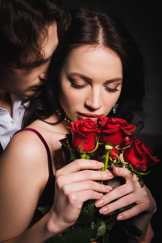 sensuale ed elegante donna che tiene rose rosse vicino all'uomo su sfondo scuro - Foto, immagini