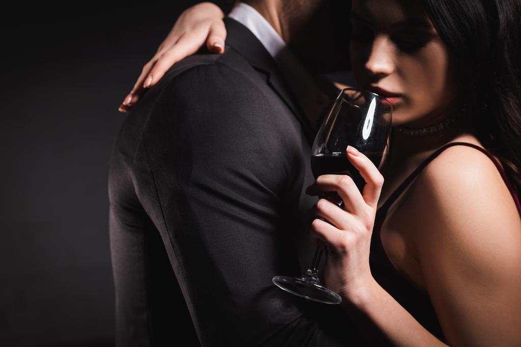 barna nő kezében pohár vörösbor miközben átölelés férfi fekete öltöny sötét háttér - Fotó, kép