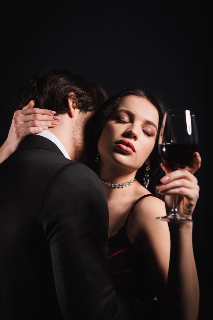sinnliche Frau mit geschlossenen Augen, die ein Glas Rotwein hält, während ein Mann im schwarzen Anzug sie isoliert auf schwarz umarmt - Foto, Bild
