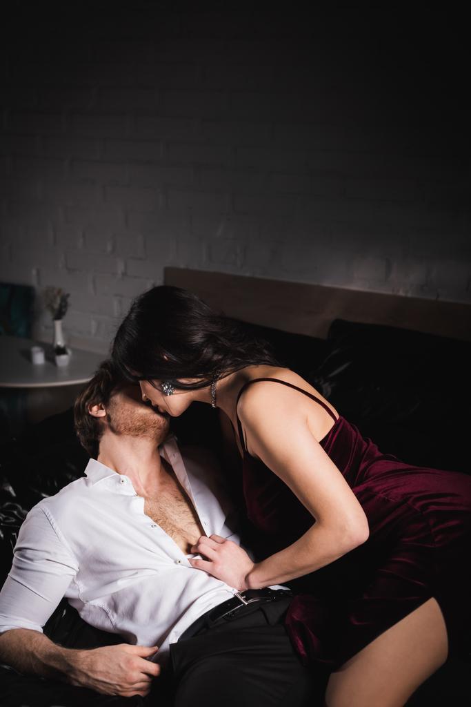 brünette Frau in elegantem Kleid küsst Mann im weißen Hemd auf schwarzem Bettzeug - Foto, Bild