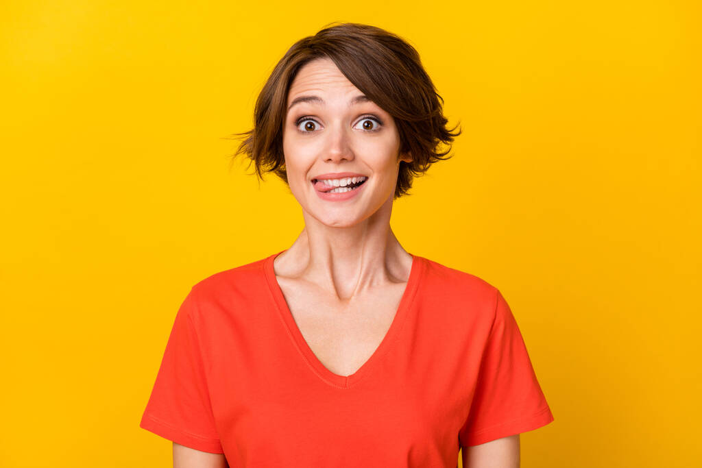 Portret van hilarisch jong meisje bijten tong uit tand glimlach kijk camera dragen oranje geïsoleerd op geel kleur achtergrond - Foto, afbeelding