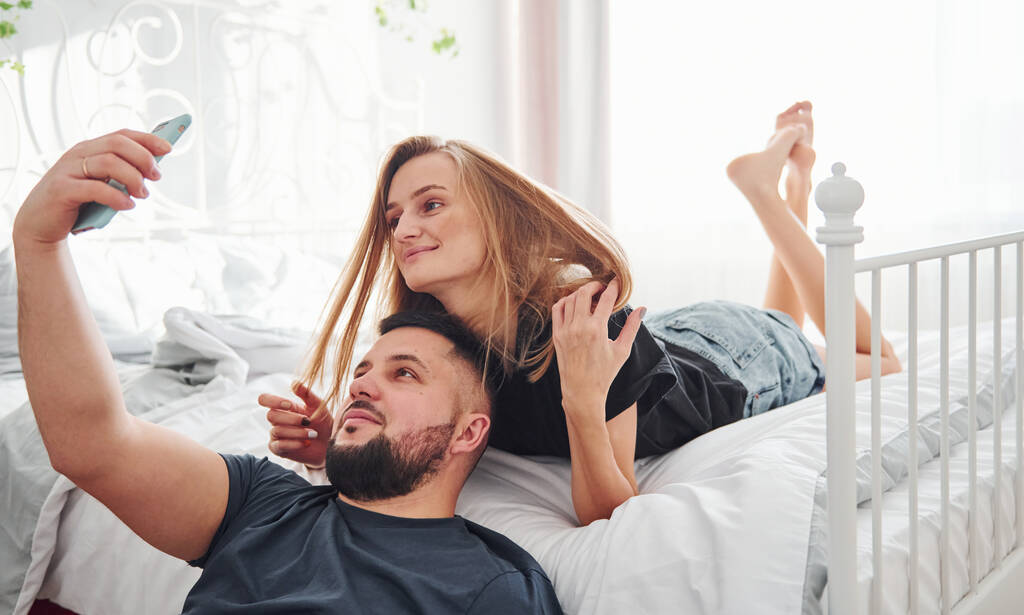 Νεαρό παντρεμένο ζευγάρι κάνει μια selfie χρησιμοποιώντας το τηλέφωνο στο υπνοδωμάτιο την ημέρα. - Φωτογραφία, εικόνα