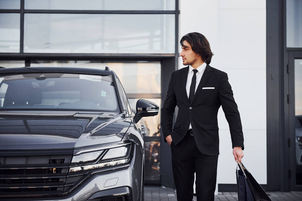 Portrét pohledného mladého podnikatele v černém obleku a kravatě venku v blízkosti moderního auta a s nákupními taškami. - Fotografie, Obrázek