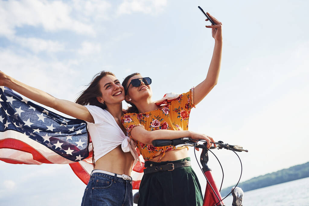 Dvě vlastenecké veselé ženy s motorkou a vlajkou USA v rukou dělá selfie. - Fotografie, Obrázek