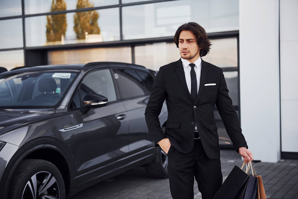 Retrato de joven hombre de negocios guapo en traje negro y corbata al aire libre cerca de coche moderno y con bolsas de compras. - Foto, Imagen