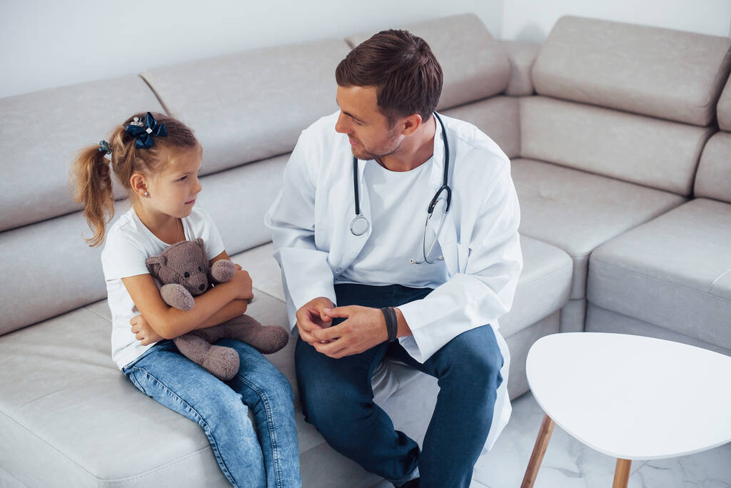 Мужчина-врач в белой форме сидит в клинике с маленькой девочкой. - Фото, изображение
