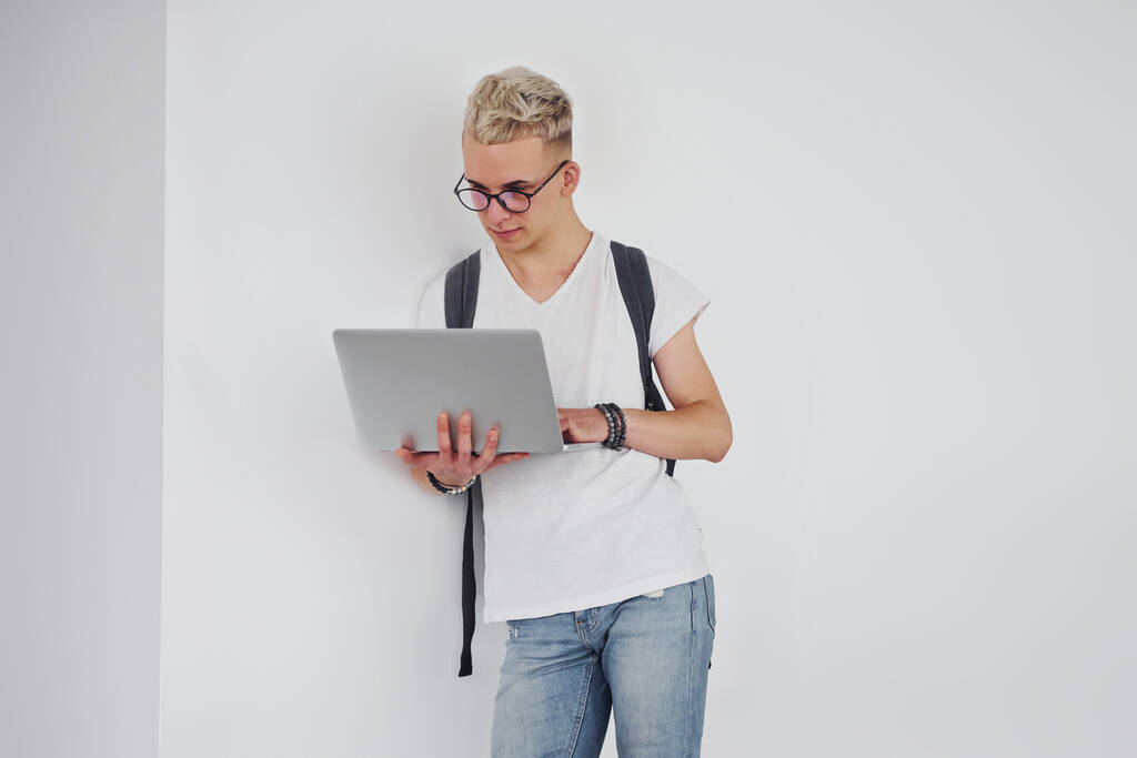 Student in casual kleding en met rugzak staat binnen tegen witte muur met laptop. - Foto, afbeelding
