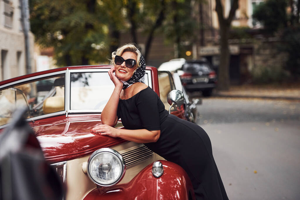 Блондинка в сонцезахисних окулярах і в чорній сукні, спираючись на старий старовинний класичний автомобіль
. - Фото, зображення