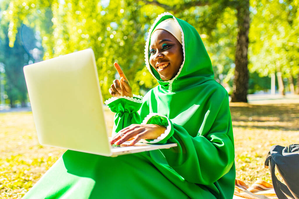 joven hermosa mujer de negocios musulmana con maquillaje moderno usar hijab verde y el uso de la computadora portátil mientras está sentado en el parque de verano - Foto, Imagen