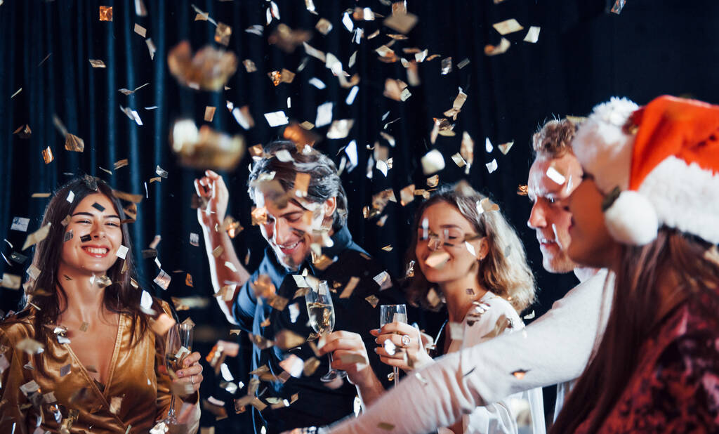 Конфетти в воздухе. Группа веселых друзей, празднующих Новый год в помещении с напитками в руках. - Фото, изображение